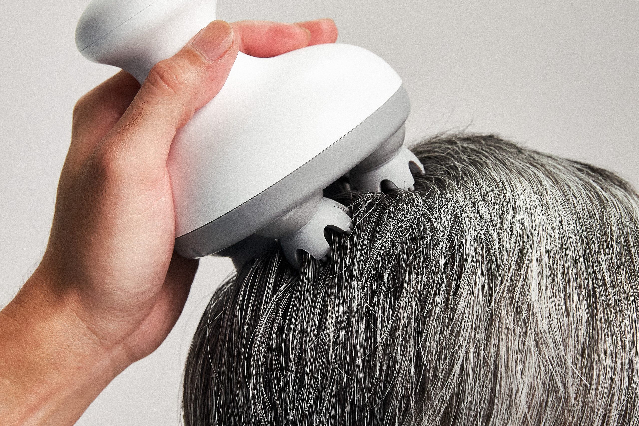 A hand using a scalp massaging device on a head of greying hair.